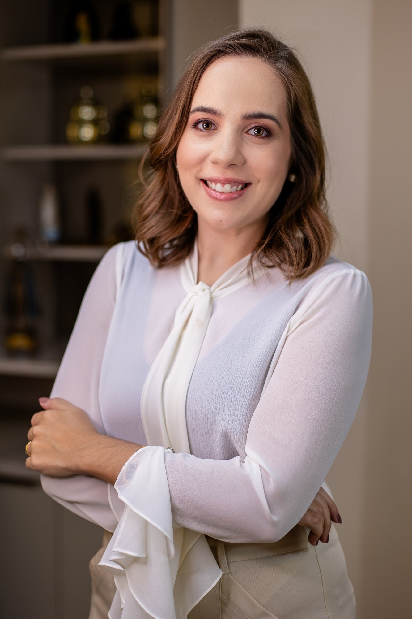 Dra. Marina Cedro sorrindo de braços cruzados, com blusa branca de laço, em seu consultório em Salvador