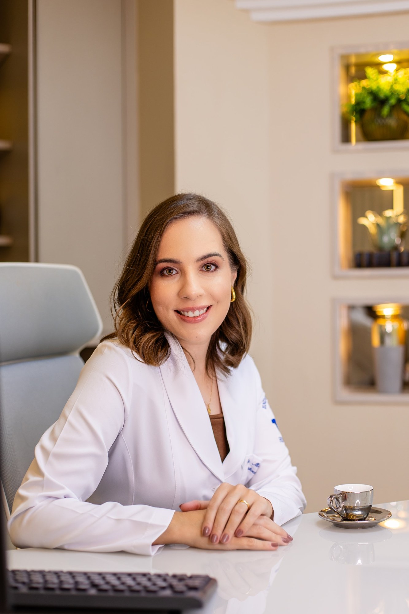 Dra. Marina Cedro de jaleco branco, sentada à mesa do consultório em Salvador, sorrindo com as mãos sobre a mesa